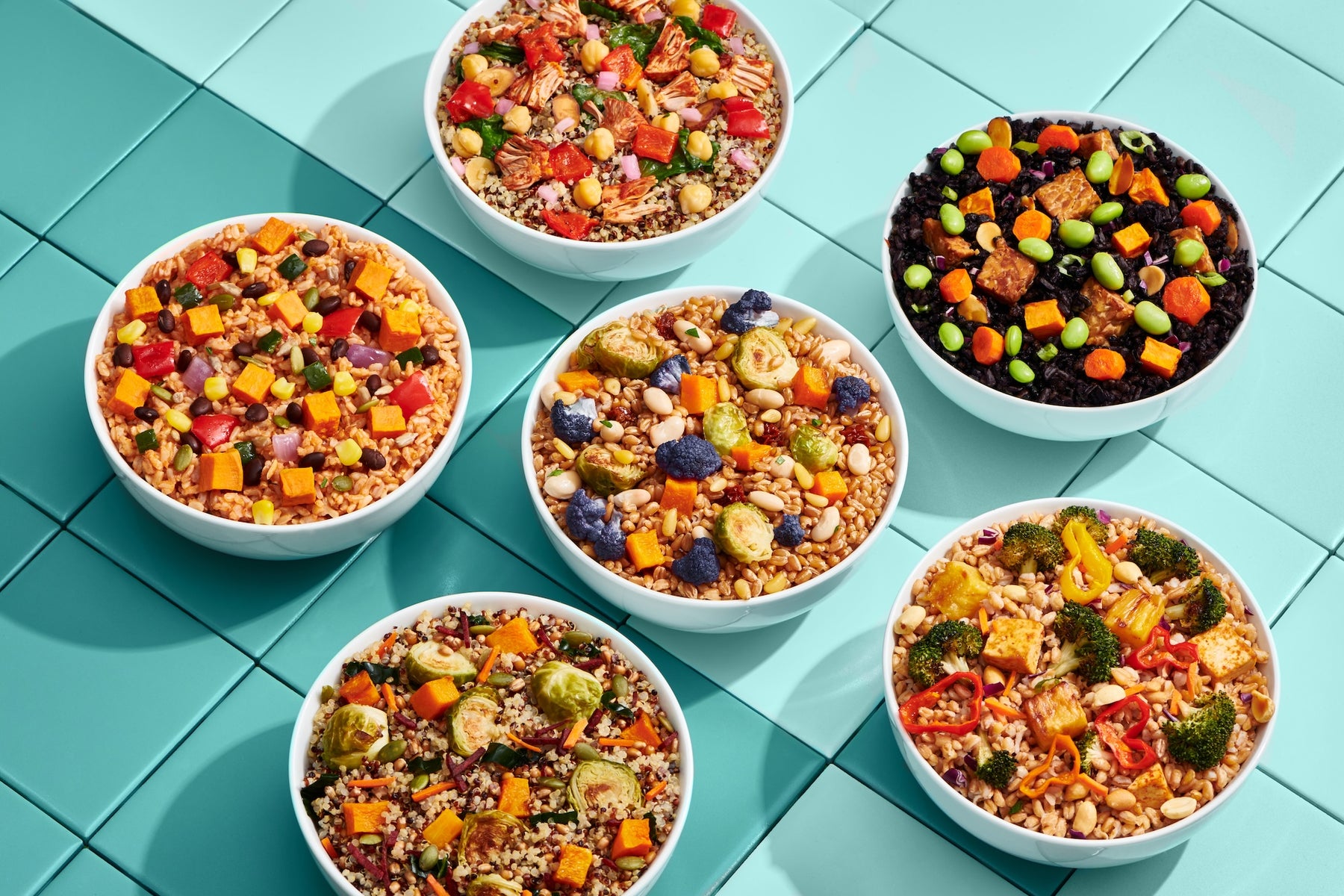 Six white bowls filled with colorful prepared grain salads arranged on a light aqua tiled surface.