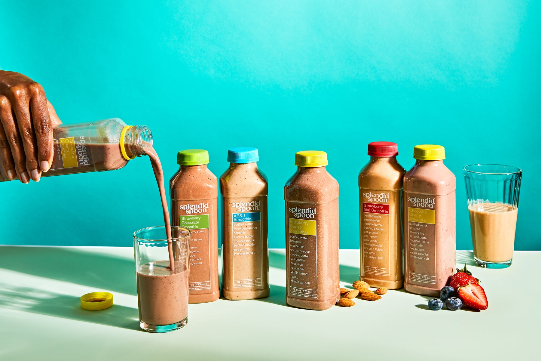 Person pouring a chocolate protein shake into a glass with protein bottles and ingredients on a teal background