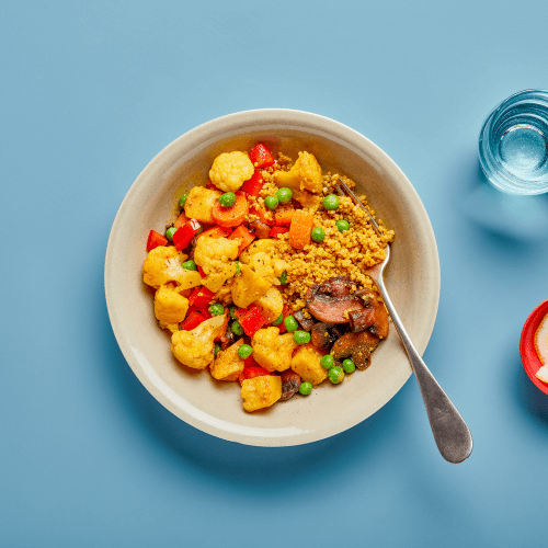 ALOO GOBI GRAIN BOWL IN A DISH ON A BLUE BACKGROUND WITH A FORK, GLASS OF WATER AND RED SALT DISH