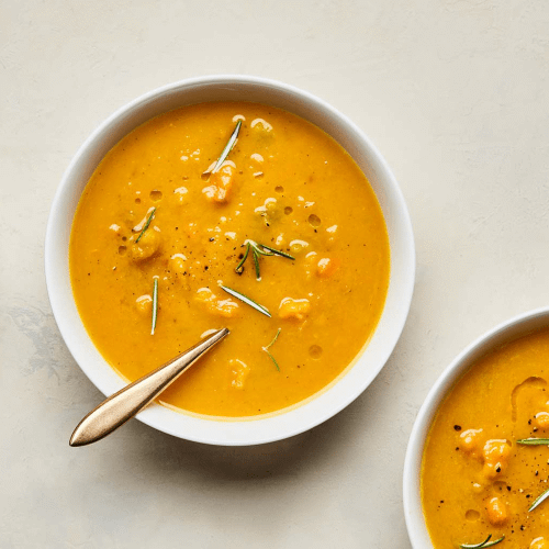 White bowl of butternut turmeric soup on marble background with gold spoon.