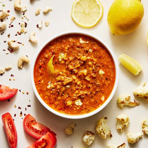 Cauliflower Tikka Soup in a white bowl surrounded by its ingredients: Tomato, Cauliflower, Lemon, and Cashews