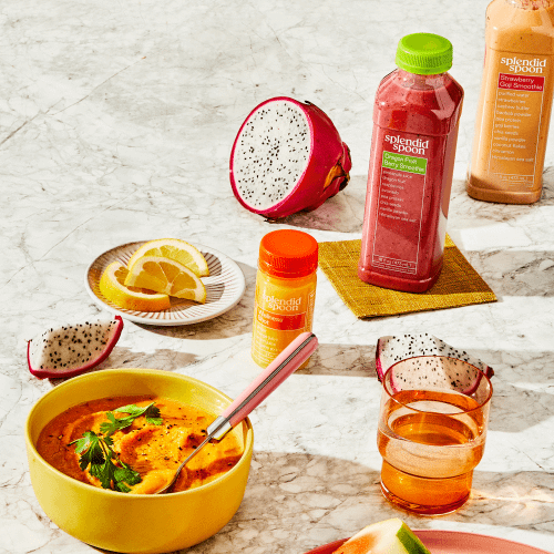 Yellow bowl of Cumin Sweet Potato Puree Soup with pink spoon, dragon fruit, sliced lemon on small dish, dragon fruit smoothie on small, yellow linen napkin, strawberry goji, wellness shot, pink, glass, clear cup, corner of a pink plate with watermelon. 
