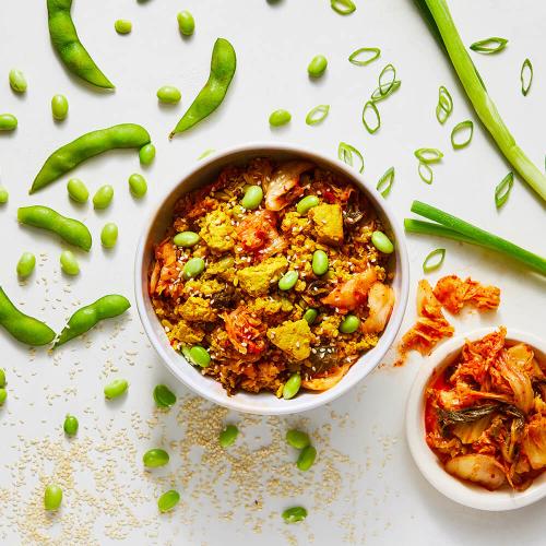 Kimchi Fried Quinoa Bowl in a white bowl surrounded by its ingredients: A bowl of kimchi, edamame in and out of the shell, and green onions