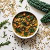 Lentil and Kale Soup in a white bowl surrounded by its ingredients: Lentils, Kale, Rosemary, and Black Pepper