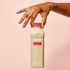 Hand holding bottle of banana flax smoothie on pink background.