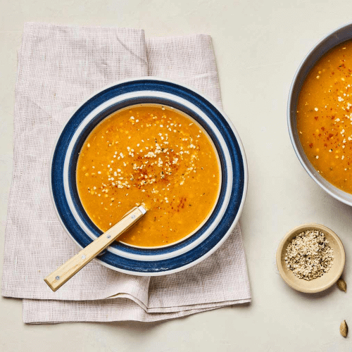 Pumpkin Pear Bisque in a blue & white rimmed bowl with a spoon on a beige linen napkin on an off-white background with a dish of hemp seeds. 