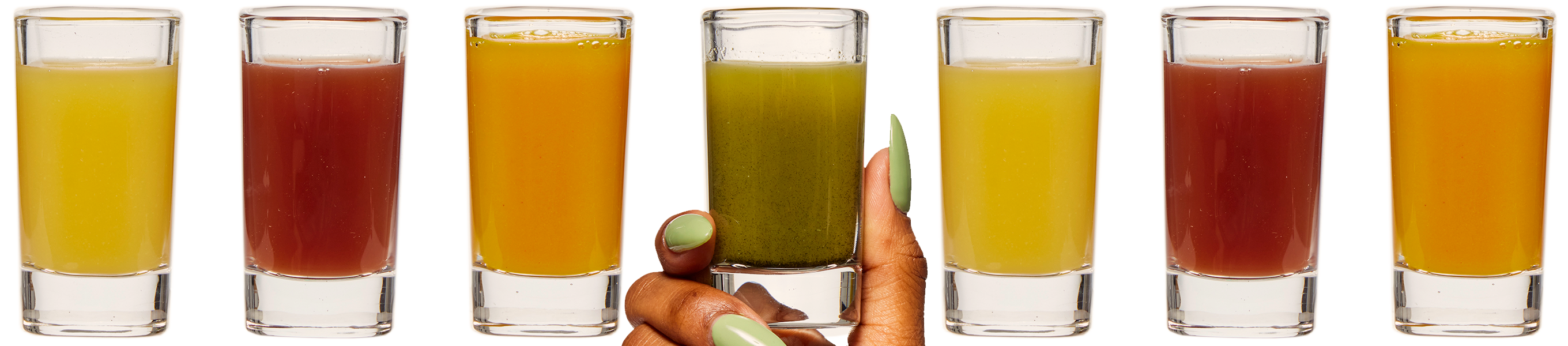 Seven shot glasses filled with colorful juices. The middle one is being held by a human hand emerging from the bottom of the image.