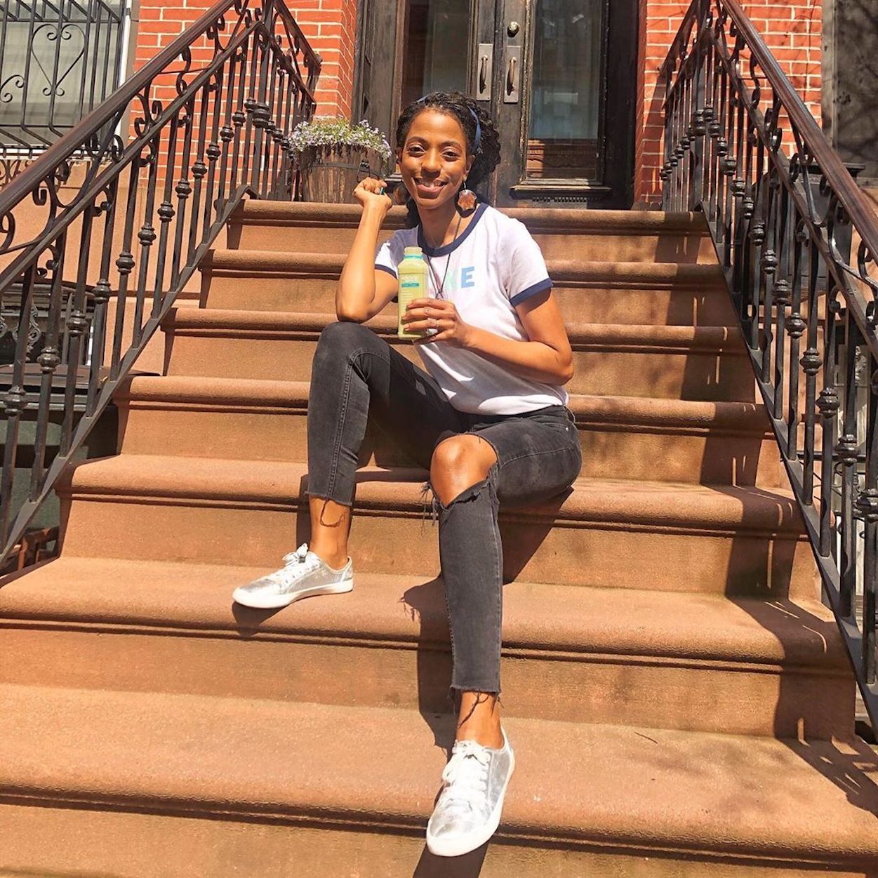 The social media personality A Brooklyn Babe sitting on the stoop of a Brownstone row house, holding a Splendid Spoon smoothie. She is sitting a a slight angle, with her feet on two different stairs.