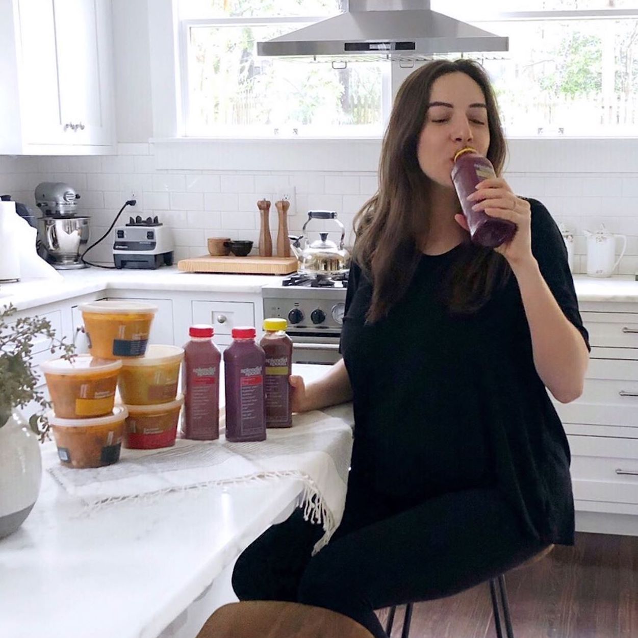 The social media personality Helene Kusman looking relaxed, sitting on a stool in a white, sun-filled kitchen, drinking a Splendid Spoon smoothie. On the counter top in front of her are a variety of Splendid Spoon smoothies and bowls.