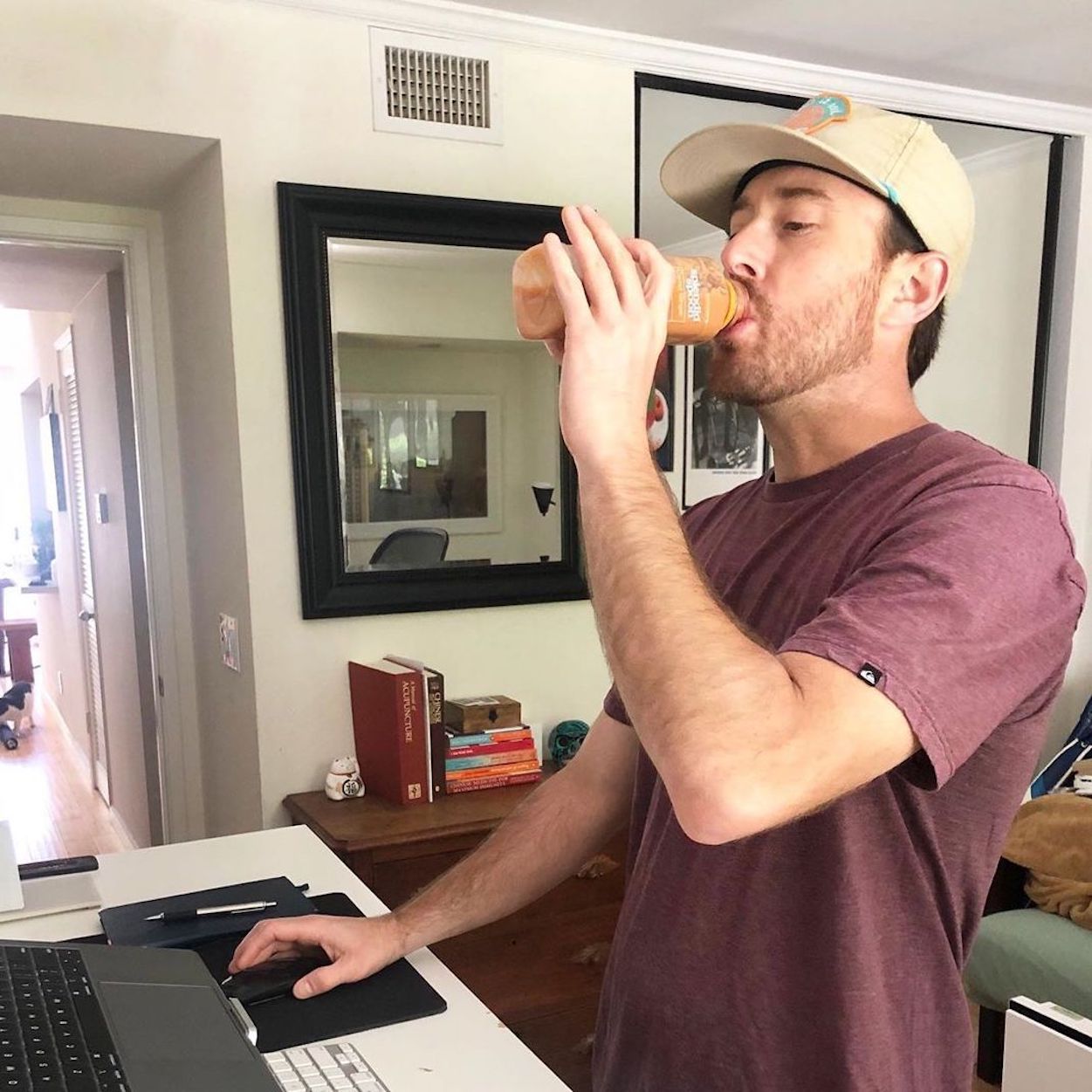 The social media personality Live to Sustain drinks a Splendid Spoon Carrot Ginger Chia smoothie in a living room. He is working on a laptop at a standing desk, and is looking at the laptop while he takes a sip. His had is on a mouse connected to the laptop.