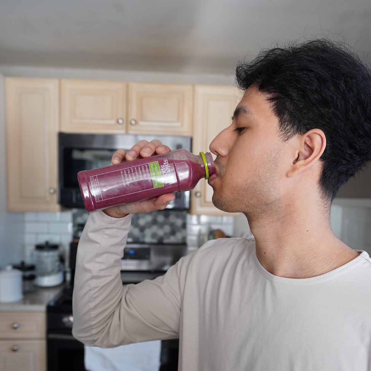 A man drinking a Splendid Spoon Dragonfruit Berry Smoothie in a kitchen, seen in profile.