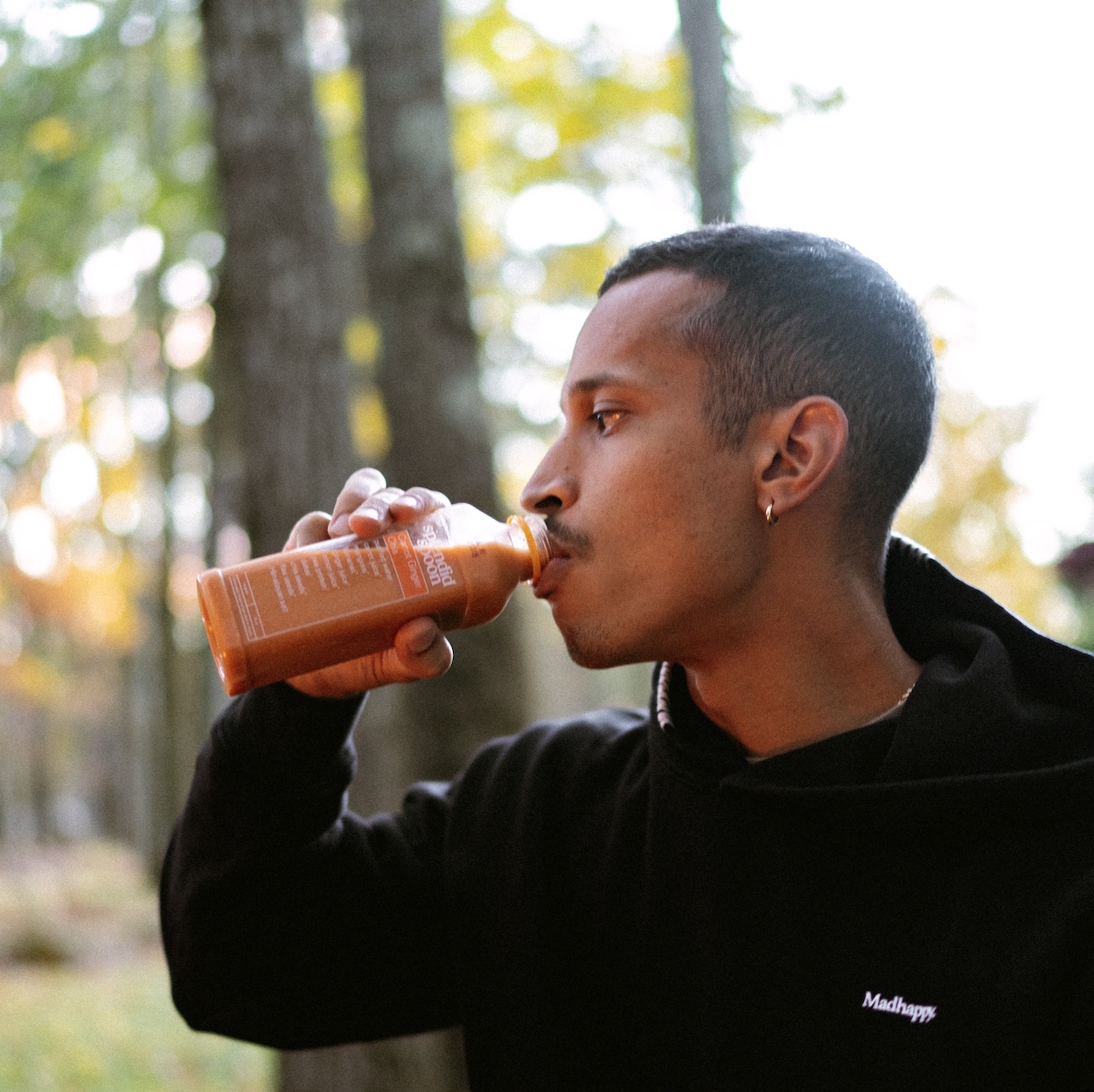 A man drinking a Splendid Spoon Carrot Ginger Chia smoothie. We see him in profile, wearing a black hoodie. Behind him are trees, with the sun is back-lighting the trees and the man's face.