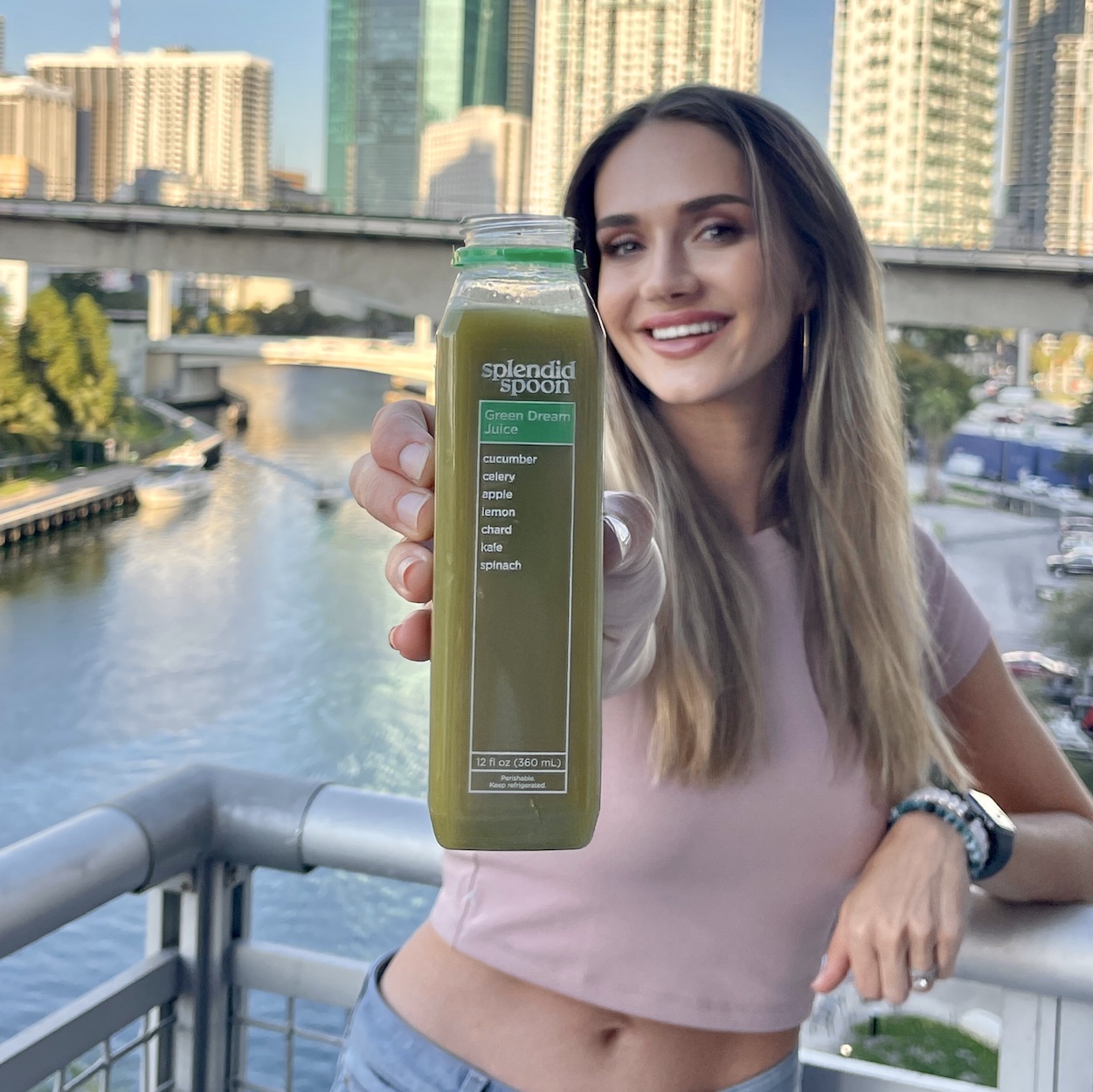 A woman stands on a balcony overlooking the Miami River in Miami, FL. Behind her are tall buildings, illuminated at sunset. She is holding a Splenid Spoon Green Dream Juice out in front of her.